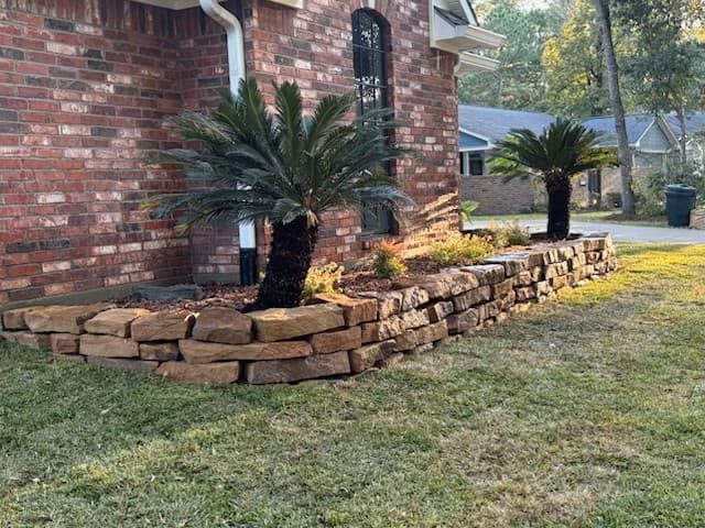 Moss rock retaining wall and planter bed with sago palms built against a brick home in Huntsville, TX